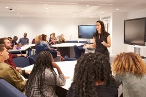 A female instructor speaks to a class of diverse students in a modern classroom setting.