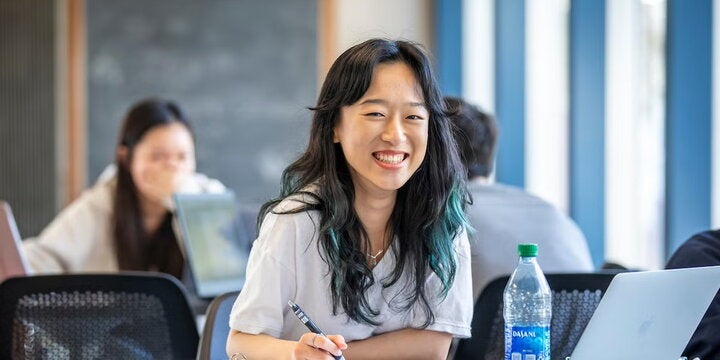A female student sits in a large study hall and looks up from her textbook.
