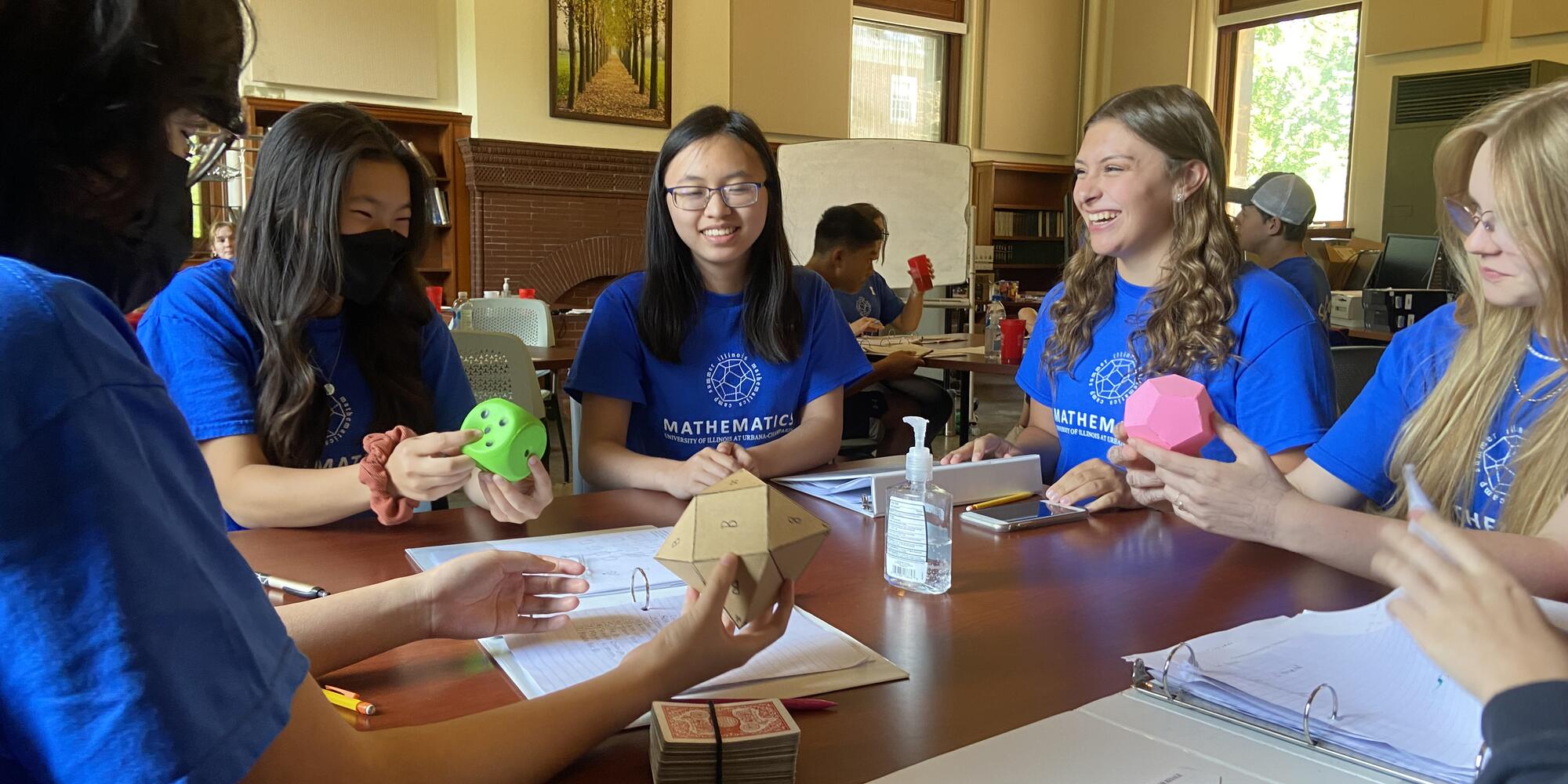 A group of students sit around a table smiling. Two are holding mathematical models. 