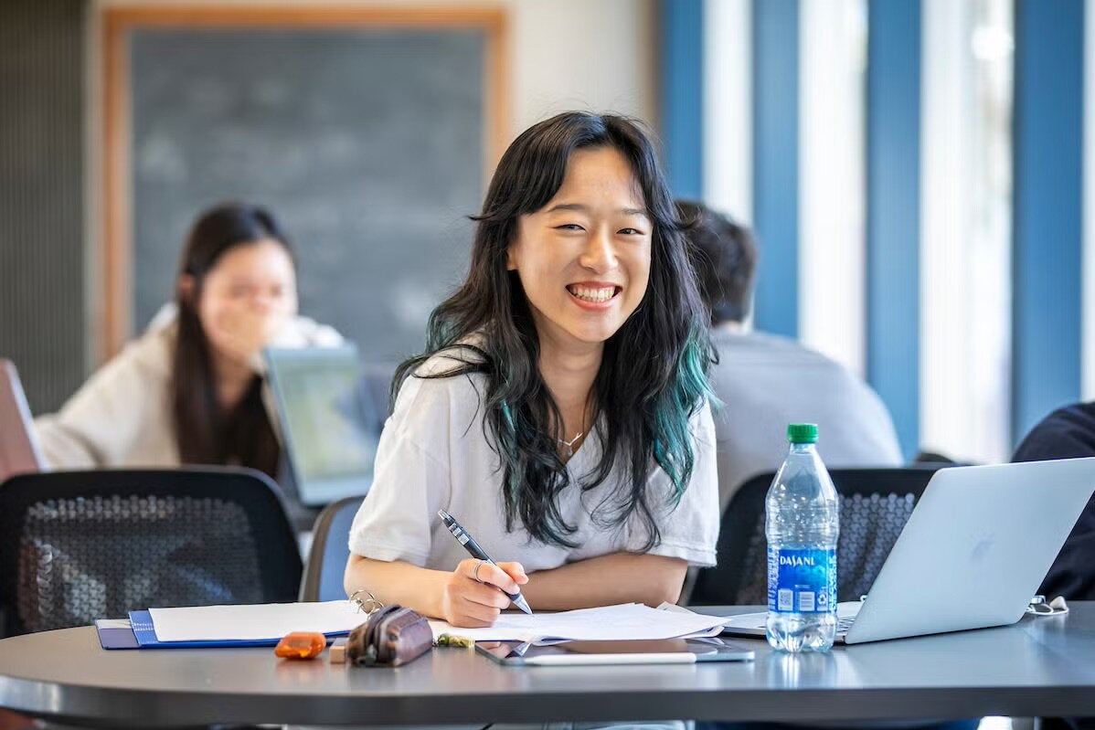 A female student sits in a large study hall and looks up from her textbook.