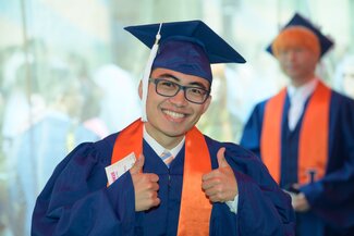 A student wearing graduation regalia gives the camera a thumbs up.