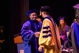 A doctoral candidate shakes the hand of his advisor. Both are wearing regalia.