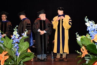 Faculty standing on stage, framed by flowers, Math Convocation 2025