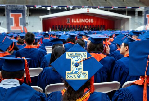 Illinois graduates sit in the audience during a commencement address. No faces are visible. In the foreground, there is a decorated graduation cap. The cap features a block I logo, representing the University of Illinois, and the "I" is filled in with lines that represent conductors or computer board circuits. 