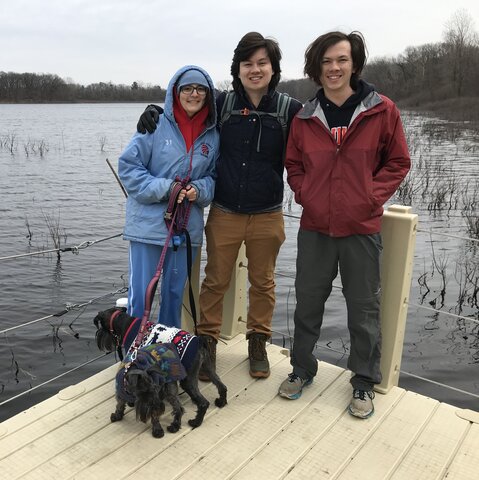 Three people wearing winter coats stand on a dock near water with two miniature schnauzers. 