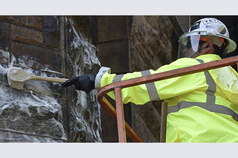 Person in hard hart and neon jacket uses a brush to clean stone on the exterior of a building.