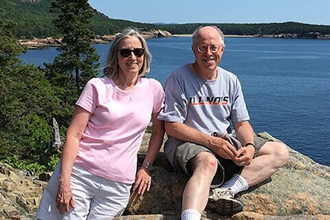 A man and woman sit on a rock in front of a large body of water. The man's shirt reads "Illinois."