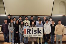 A group of students stand in front of a blackboard. A few students in the front are holding a sign that says "IRisk: Illinois Risk Lab."