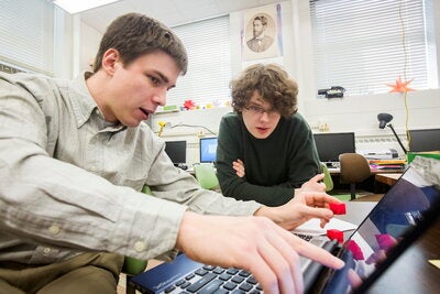 Two students sit at a table, collaborating on a project for the Illinois Geometry Lab. One student points at a laptop screen and the other student looks to see what they are pointing at.