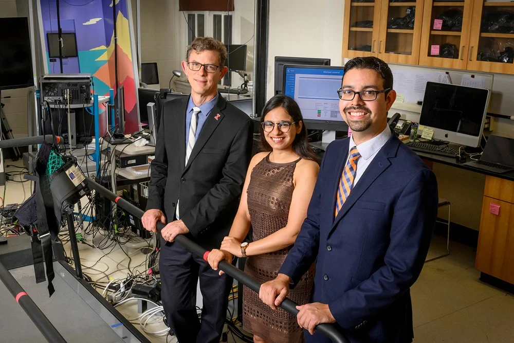 Richard Sowers, Rachneet Kaur and Manuel Hernandez stand next to treadmill in a lab. They are surrounded my monitors, electrical cords, and medical monitoring equipment
