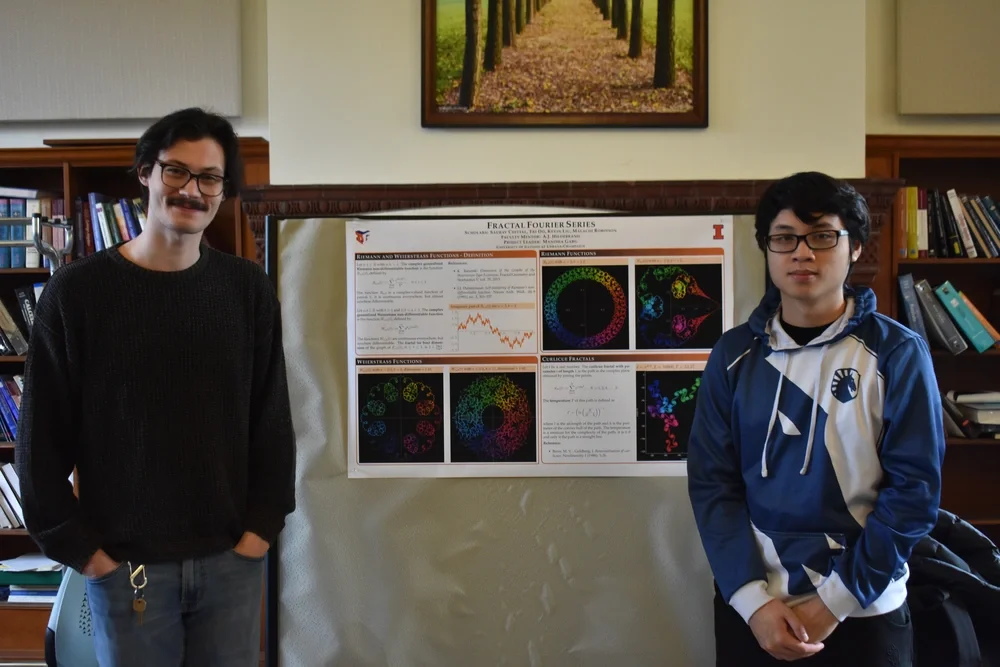 Two students stand in front of their research poster. The poster contains colorful fractal patterns on a black background.