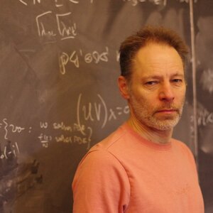 A man stands in front of a chalkboard. There are mathematical equations on the chalkboard.
